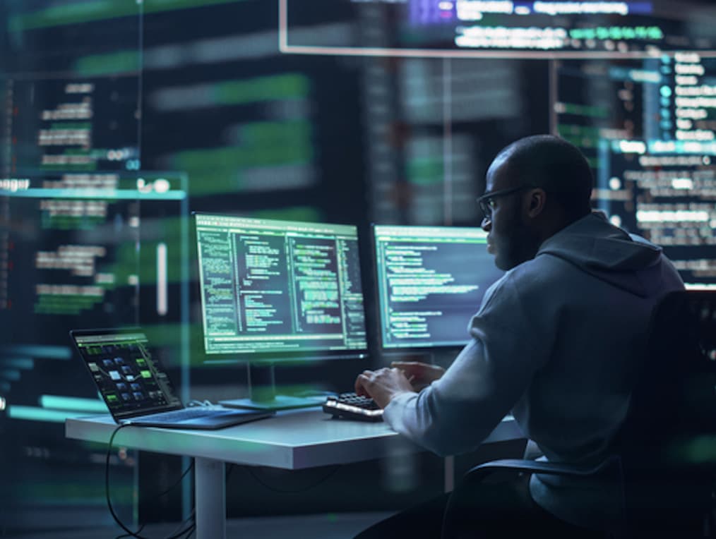 An engineer, looking on monitors with a laptop on the desk.